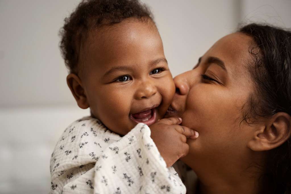 mãe beijando seu filho que sorri mostrando os primeiros dentinhos.